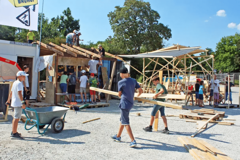 Kinder schieben Schubkarren und tragen Holz, um ein kleines Haus zu bauen.