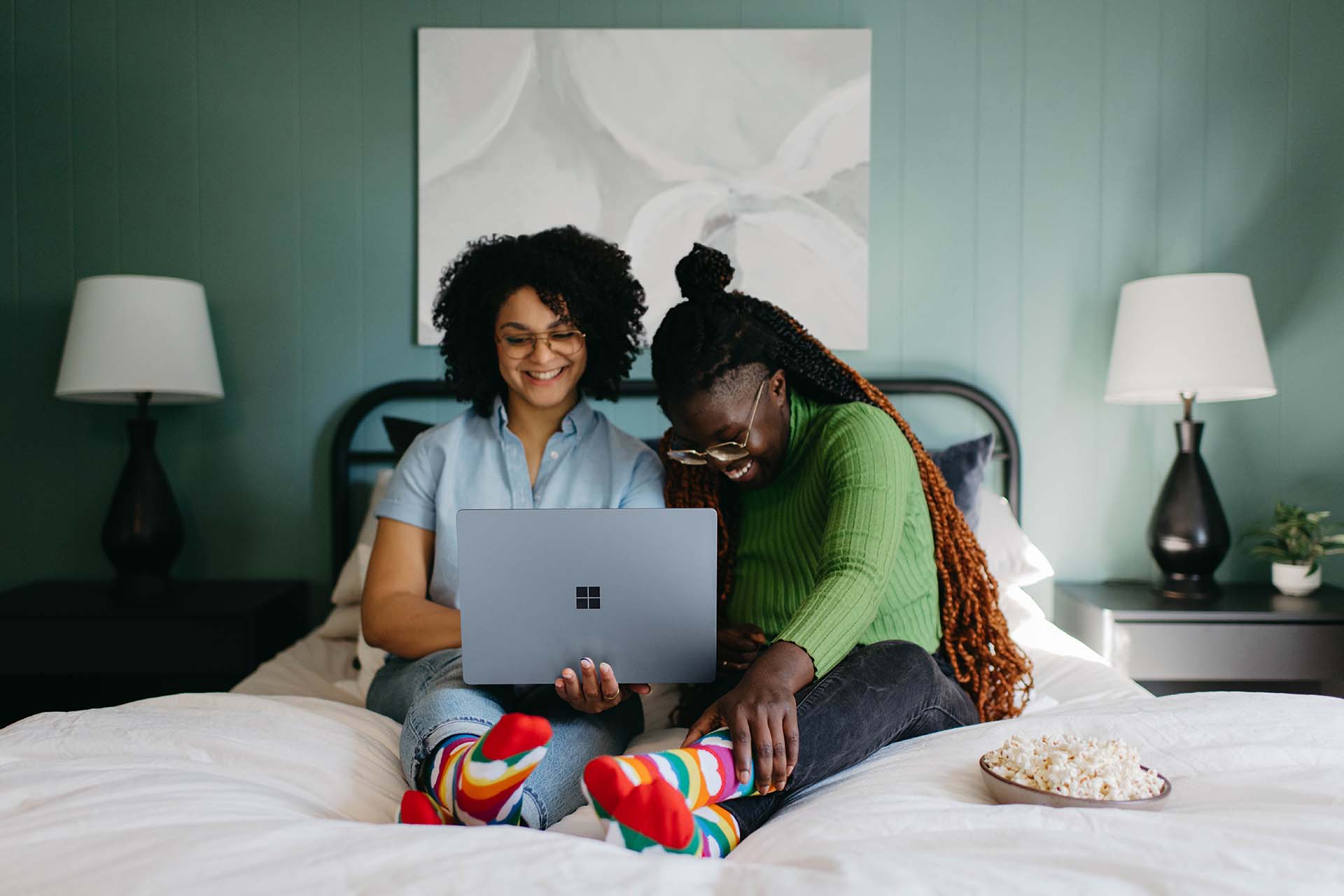 Zwei Frauen sitzen auf einem Bett und blicken fröhlich in ein Laptop