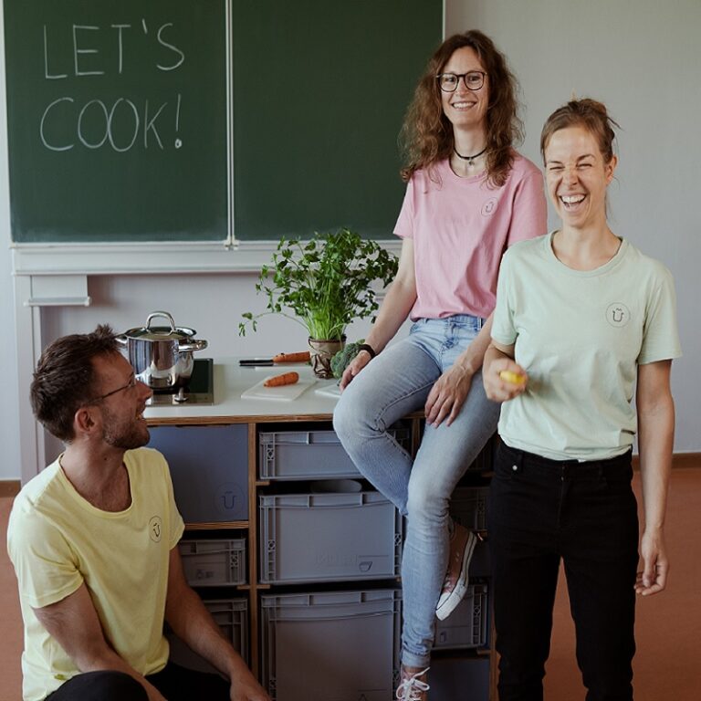 Drei Menschen lachend an einer Mobilen Küche in einem Klassenzimmer
